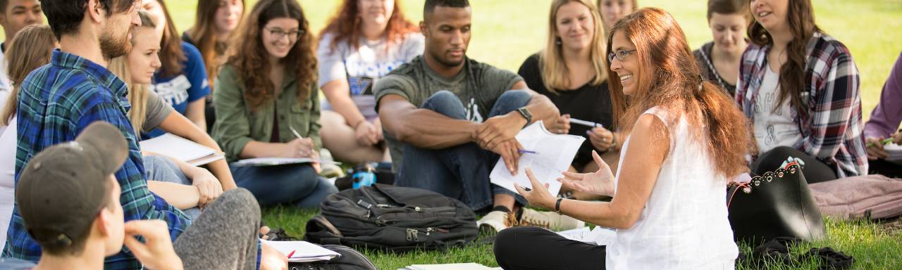 Dr. Val Peterson sitting on the lawn with students for class, smiling.